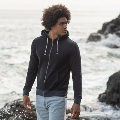 Man wearing the recycled contrast zip hoodie in Black/ Charcoal on a rocky shoreline