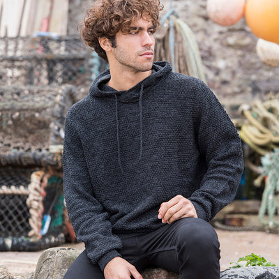 Man wearing the knitted recycled hoodie in Charcoal/ Black, seated by lobster pots