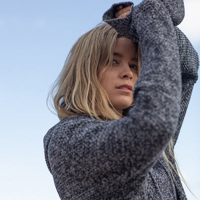 Close-up of a woman wearing the knitted recycled hoodie, arm raised against a blue sky