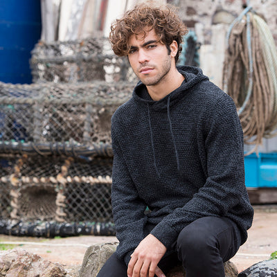 Man wearing the knitted recycled hoodie in Charcoal/ Black, seated by lobster pots