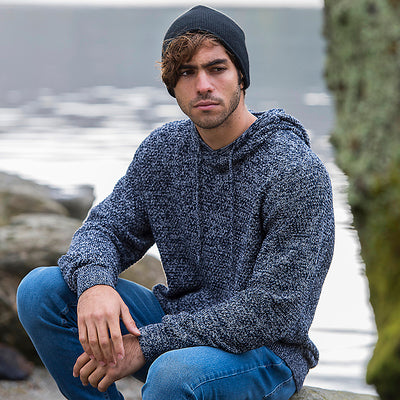 Man in a beanie wearing the knitted recycled hoodie in Navy/ Heather Grey, sitting by a lake